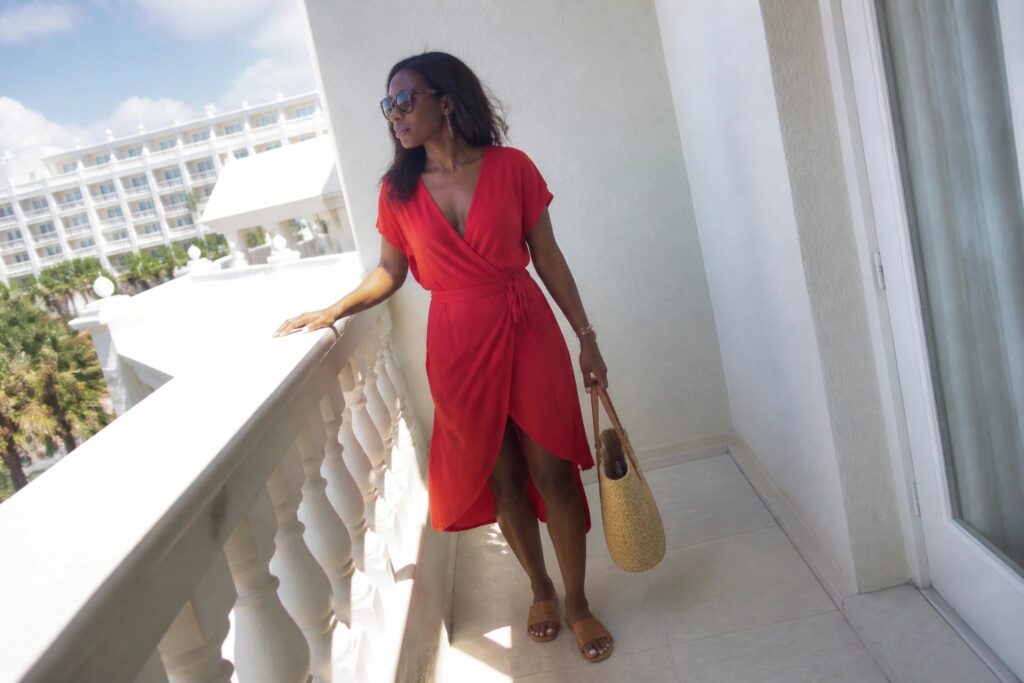 Woman in red wrap dress holding straw tote on sunny oceanfront balcony