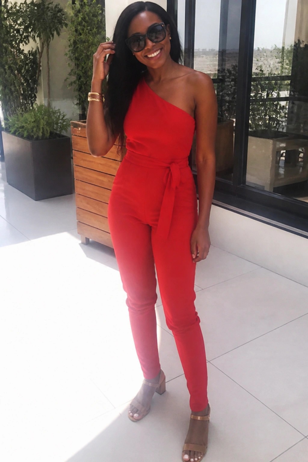Woman in red one-shoulder jumpsuit smiles outdoors wearing sunglasses and heels.