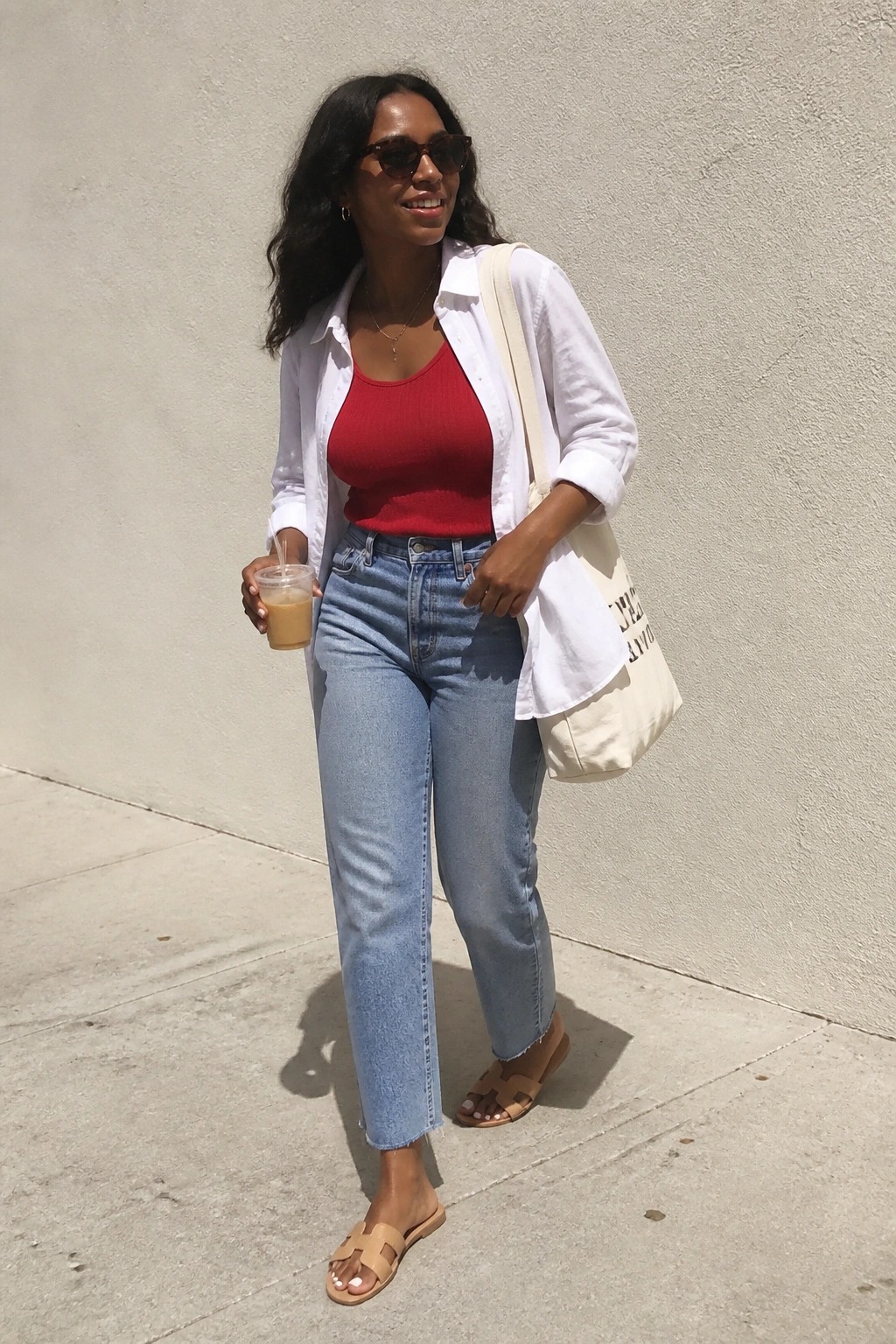 Smiling woman in red tank top, open white shirt, blue jeans, holding drink and tote.