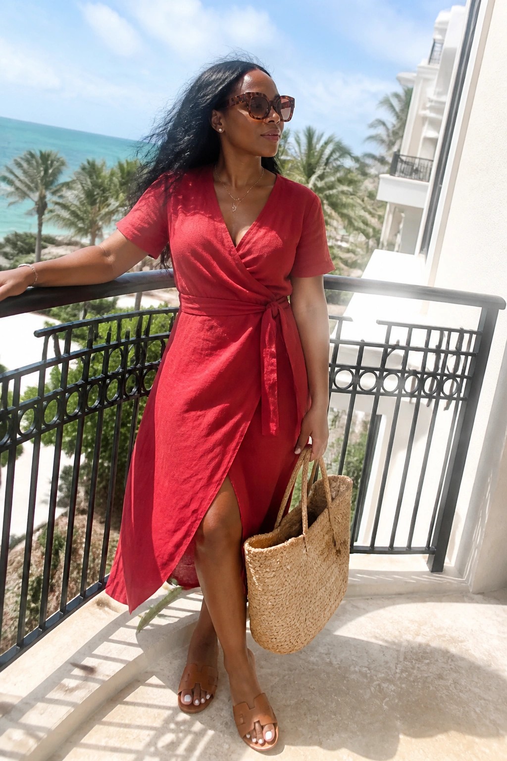 Woman in red wrap dress holding straw tote on sunny oceanfront balcony