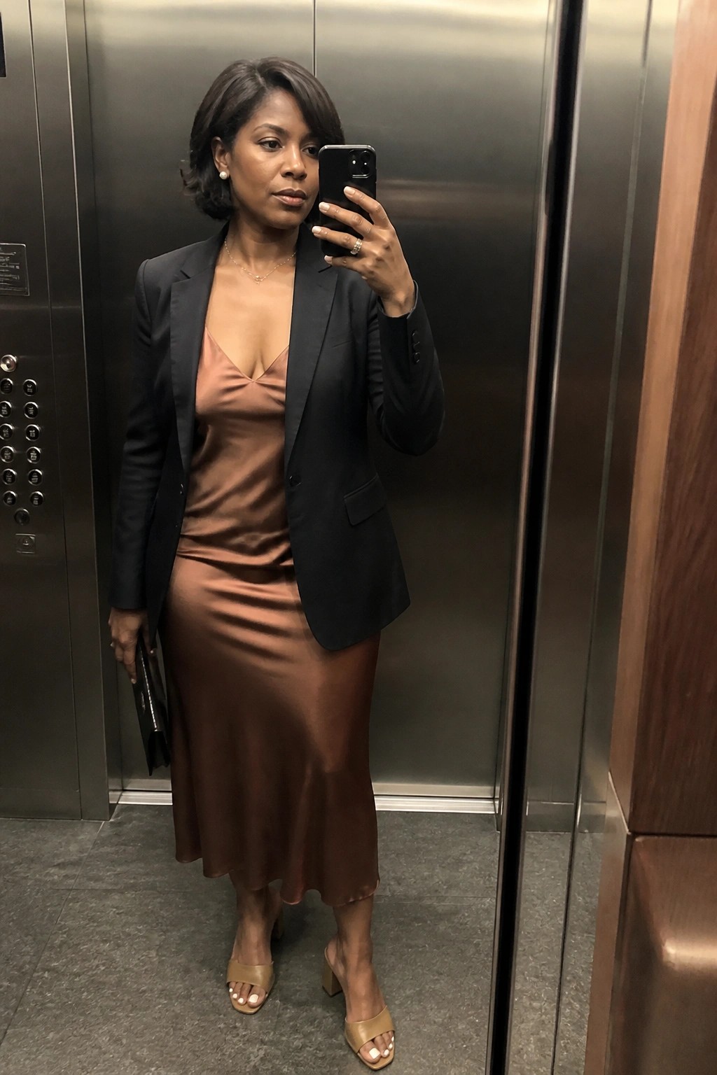 Woman in brown satin dress and black blazer taking selfie in elevator