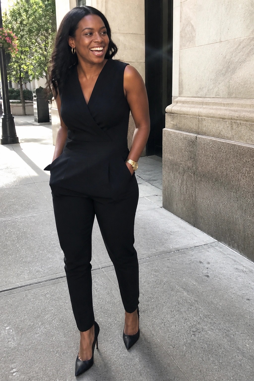 Smiling woman in black sleeveless jumpsuit and heels by stone building