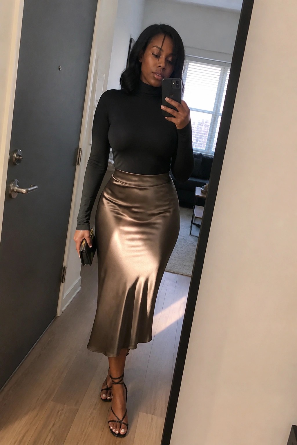 A Black woman wearing a fitted black long sleeve turtleneck top tucked into a bronze metallic satin midi skirt paired with black strappy high heels and holding a small black clutch while taking a mirror selfie.