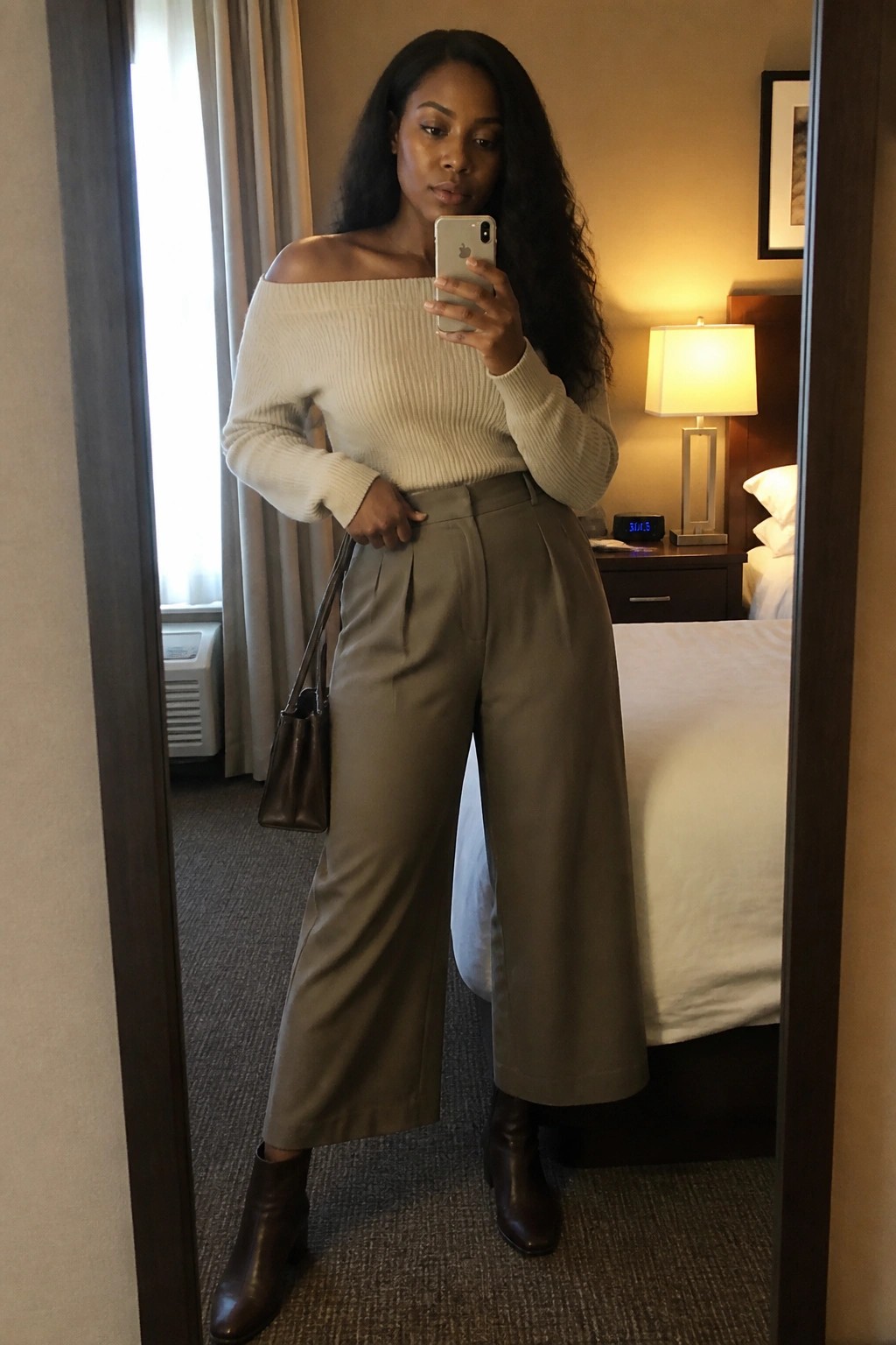 Woman in off-shoulder cream sweater and wide-leg pants taking mirror selfie in hotel room