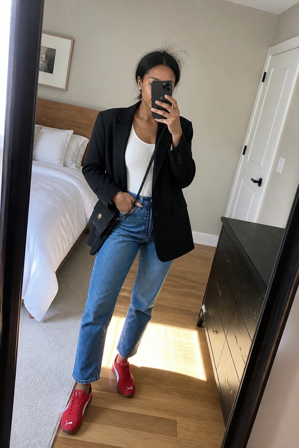 Woman in black blazer, white tank top, blue jeans, and red sneakers in bedroom mirror selfie