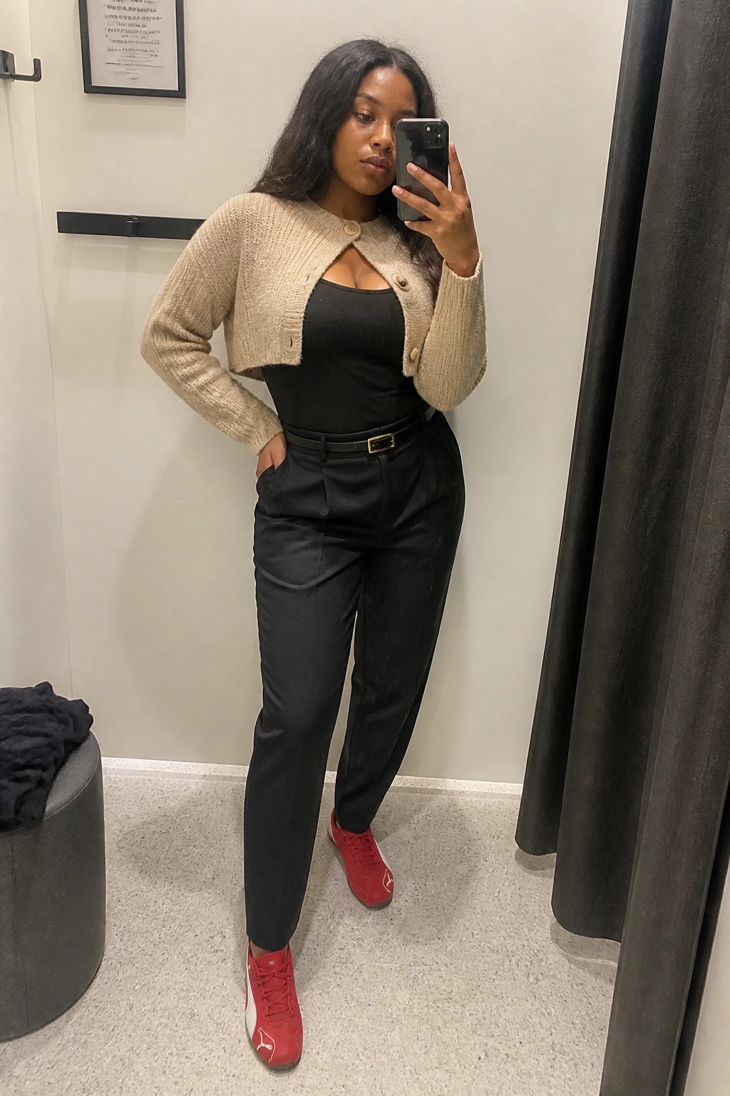 Woman in a beige cropped knit cardigan over black top and high waisted black pants with belt, paired with red Puma sneakers, posing for a mirror selfie indoors.