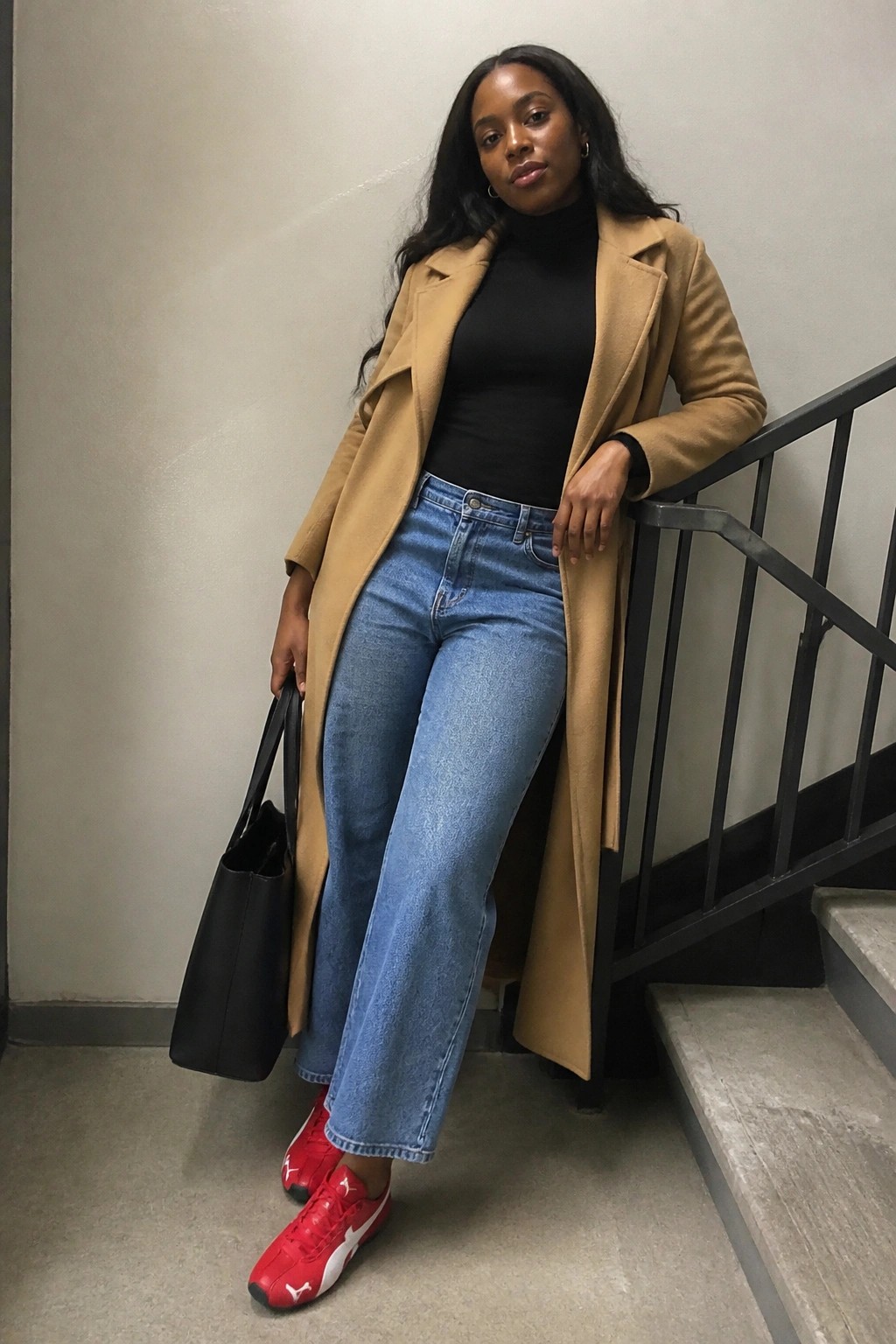 Woman in tan coat, black turtleneck, blue jeans, red sneakers on stairs with black bag