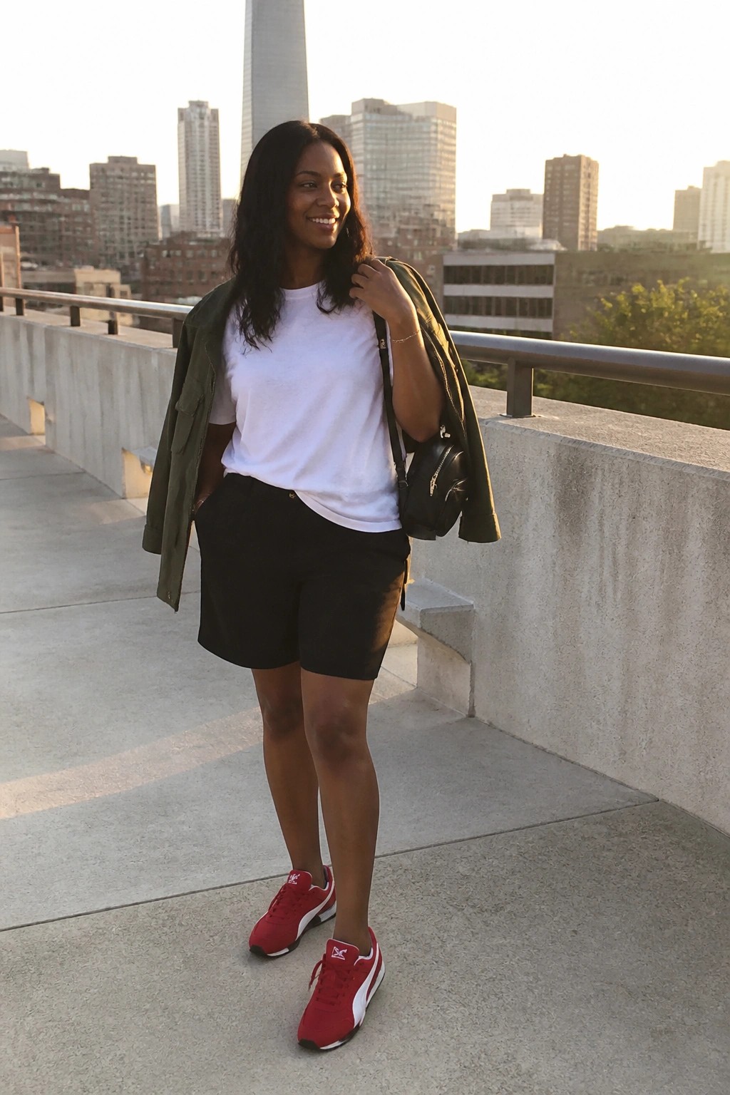 Smiling woman in white t-shirt, black shorts, red sneakers, green jacket over shoulder, city skyline.