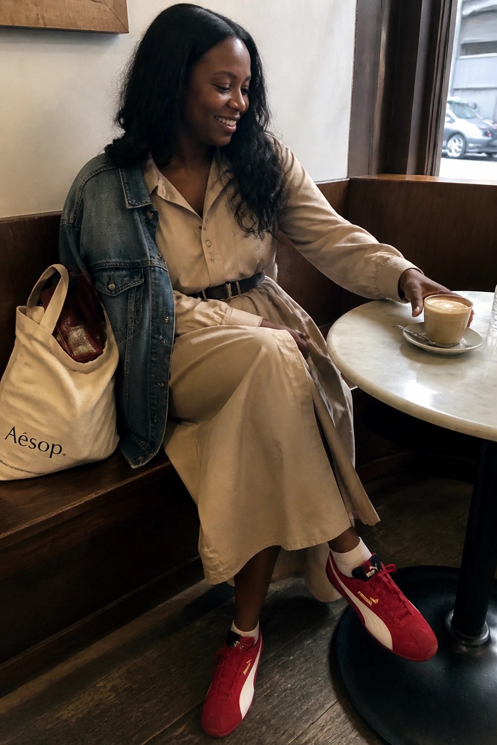 Woman in beige dress and denim jacket reaches for coffee wearing red Puma sneakers