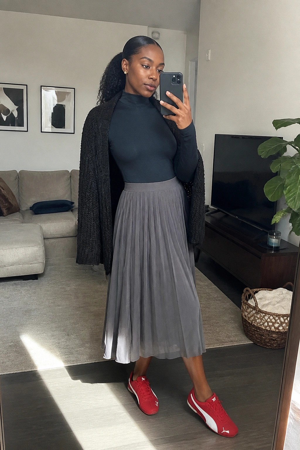 Woman in gray turtleneck, pleated skirt, black cardigan, and red sneakers taking mirror selfie.