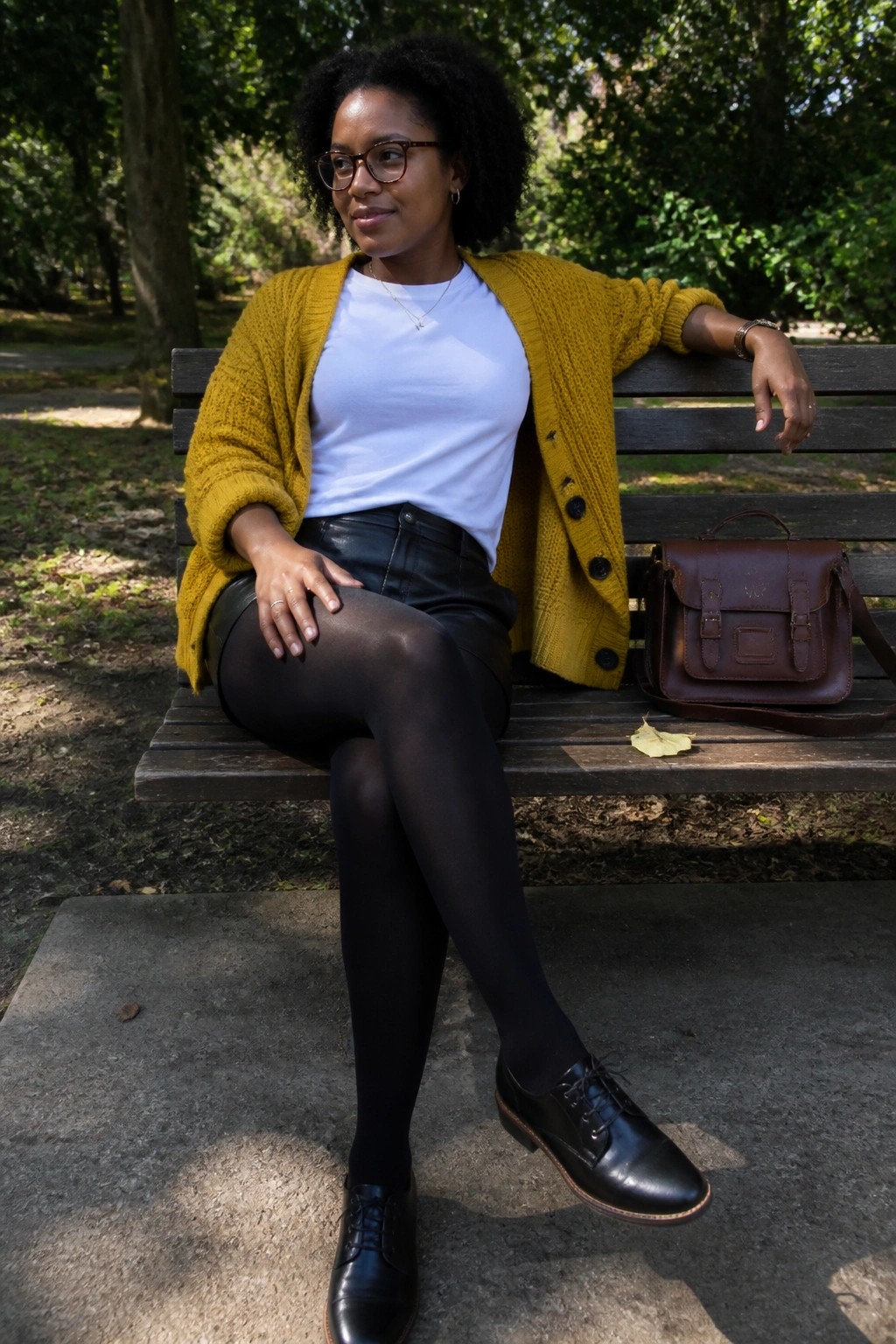Woman in yellow cardigan and black tights sits on park bench with satchel