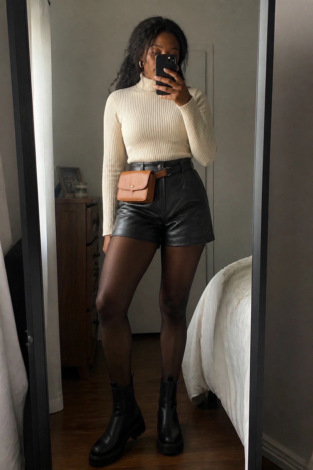 Woman in cream turtleneck and black leather shorts taking mirror selfie with brown belt bag