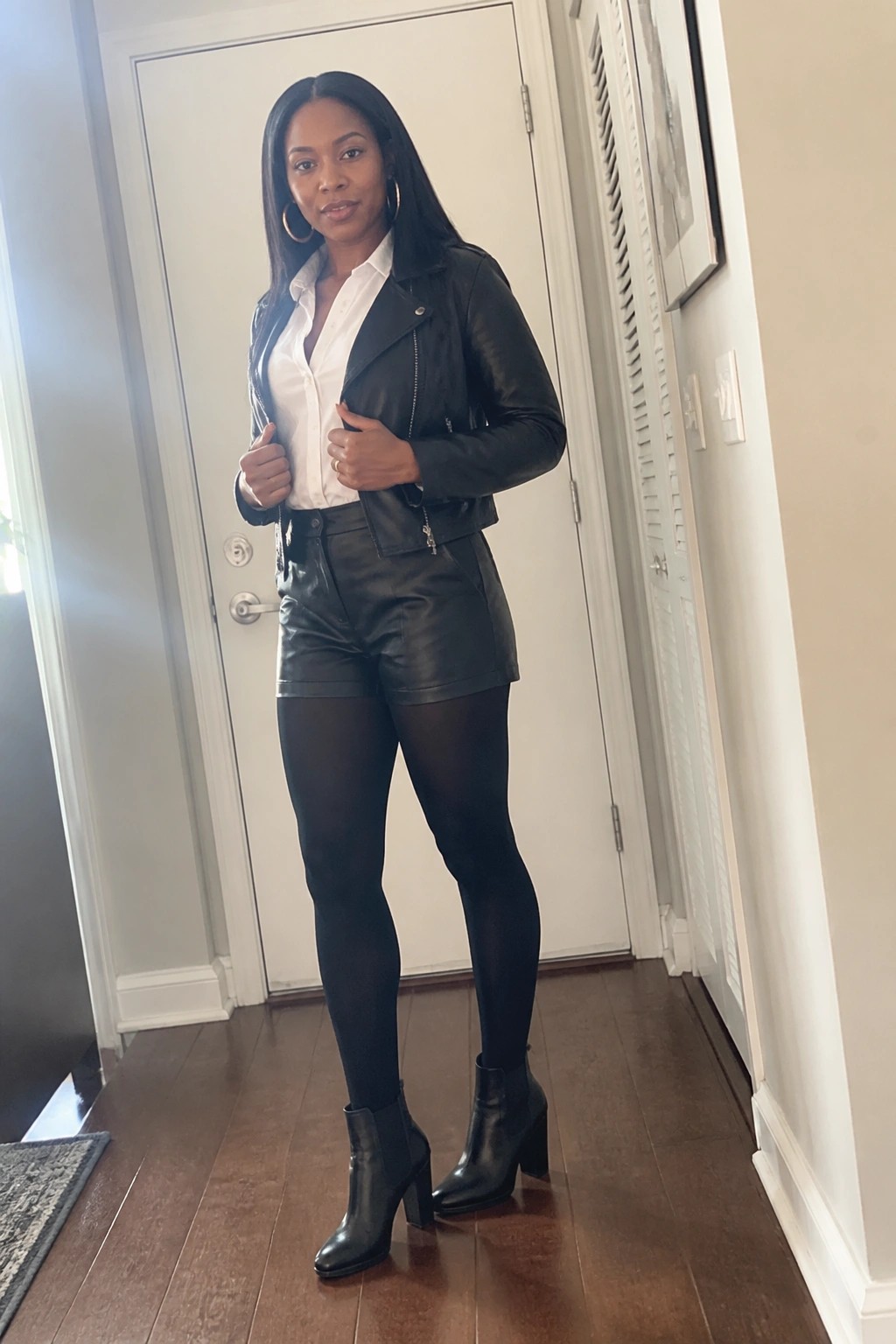 Woman in black leather jacket, white shirt, shorts, tights, and boots in hallway
