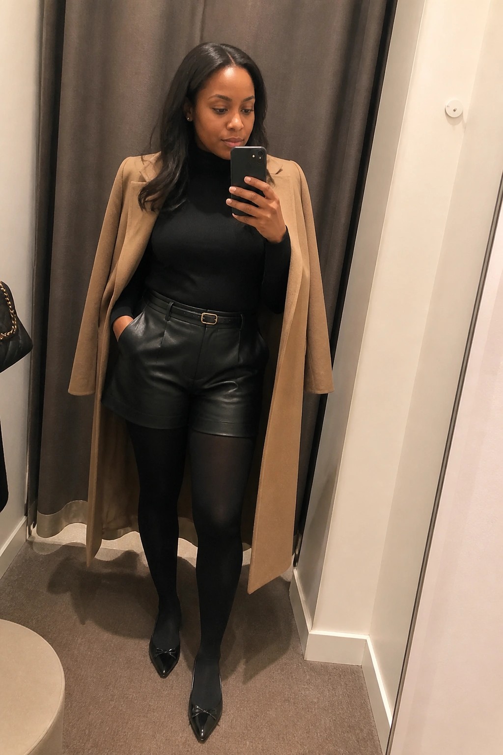 Woman in black turtleneck and leather shorts with open tan coat taking mirror selfie