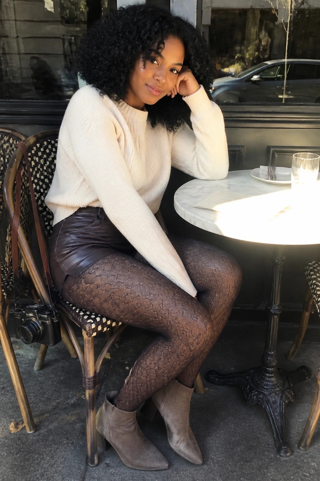 Woman with curly hair in white sweater, leather shorts, and patterned tights at outdoor cafe.