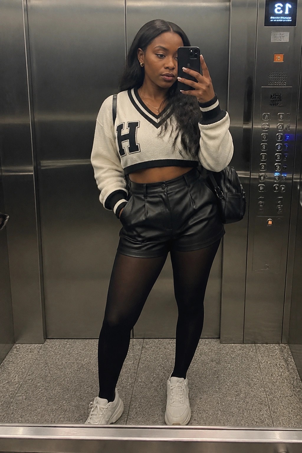 Woman in cropped varsity sweater and leather shorts taking selfie in elevator