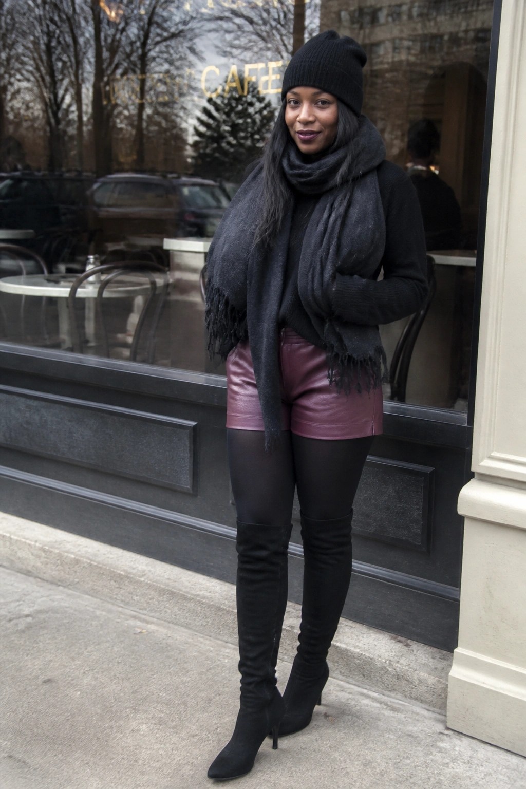 Woman in black beanie, scarf, and thigh-high boots with maroon leather shorts by cafe
