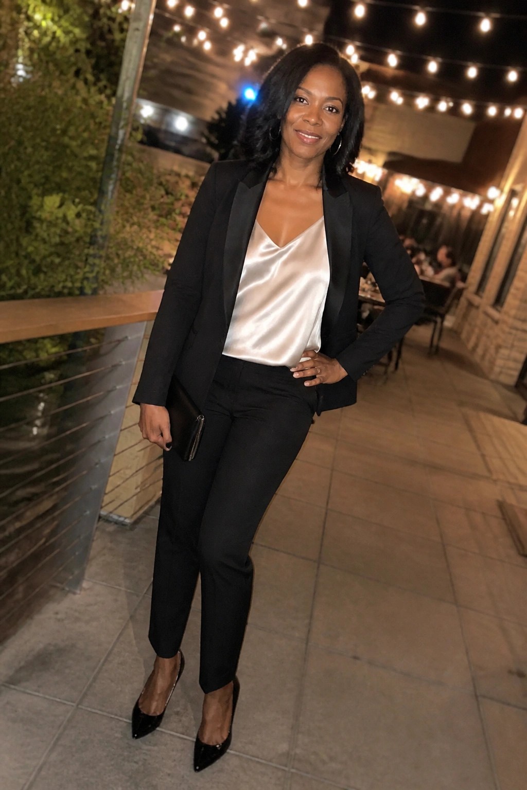 Woman in black blazer over white satin top with black pants at night patio
