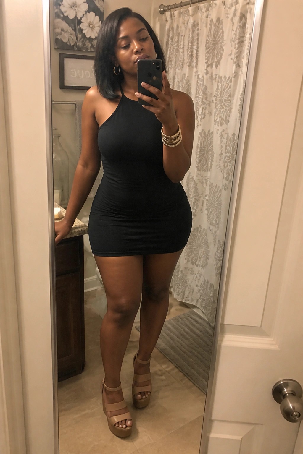 Woman in black halter mini dress taking mirror selfie in bathroom wearing beige heels.
