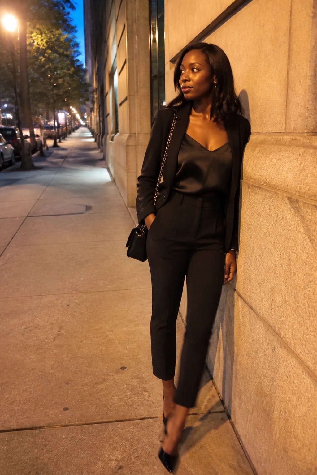 Woman in black suit leans against stone wall on nighttime city sidewalk.