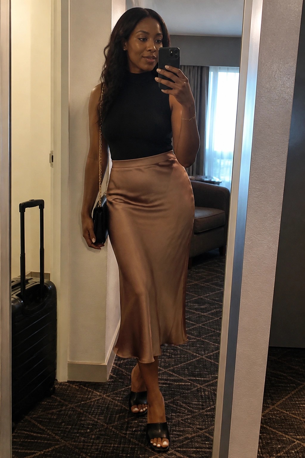 Woman in black top and satin skirt takes mirror selfie in hotel with suitcase nearby