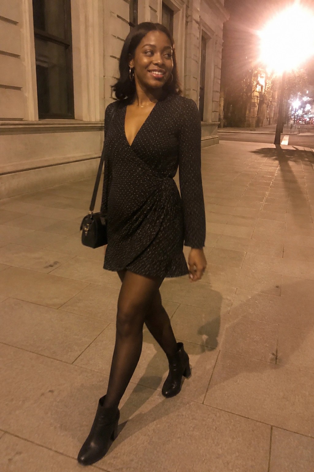 Woman in black sparkly wrap dress with handbag walks on nighttime sidewalk