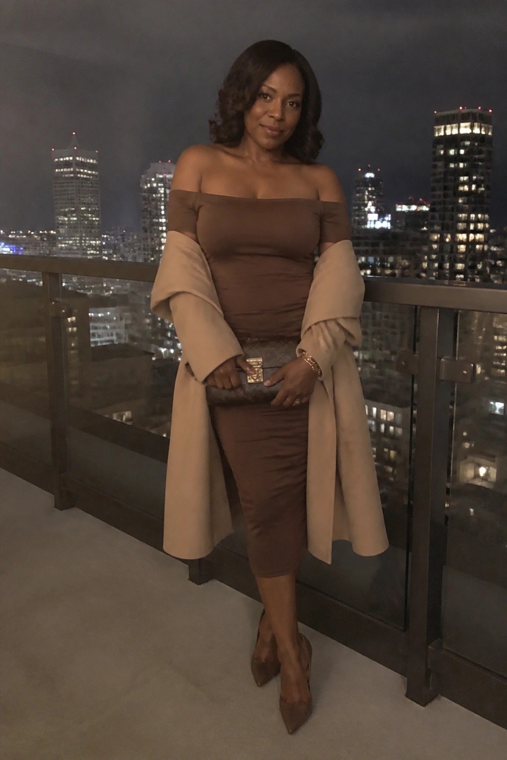 Stylish woman in brown off-shoulder dress and beige coat holding clutch on city balcony at night.