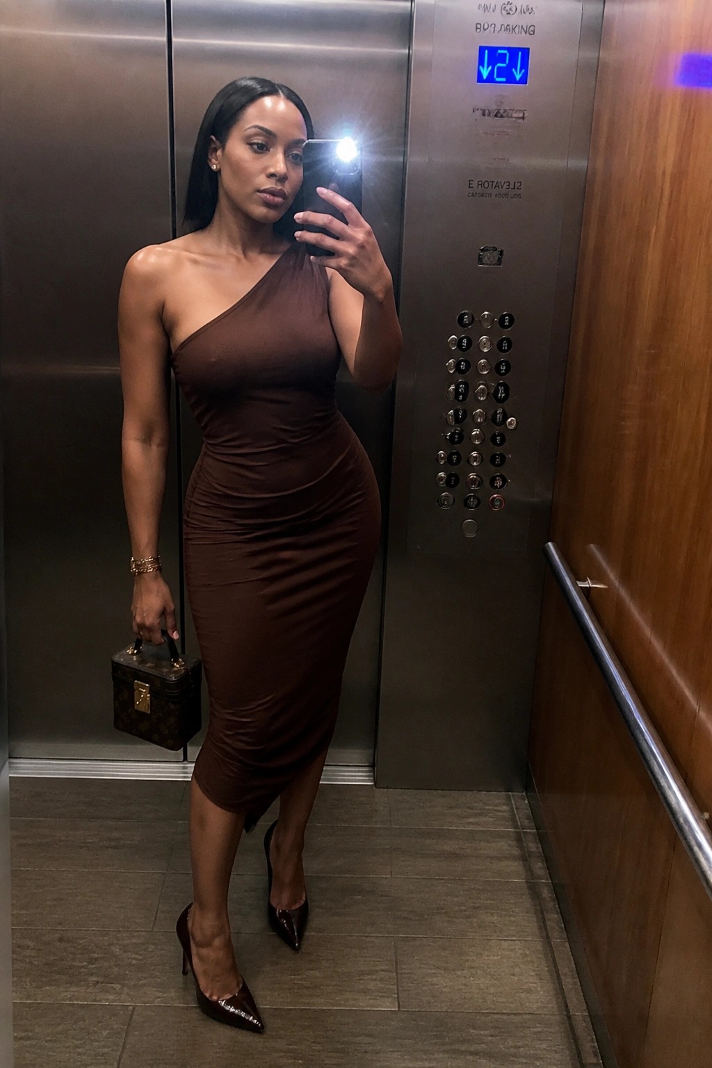 Woman in brown one-shoulder dress taking selfie in elevator with handbag