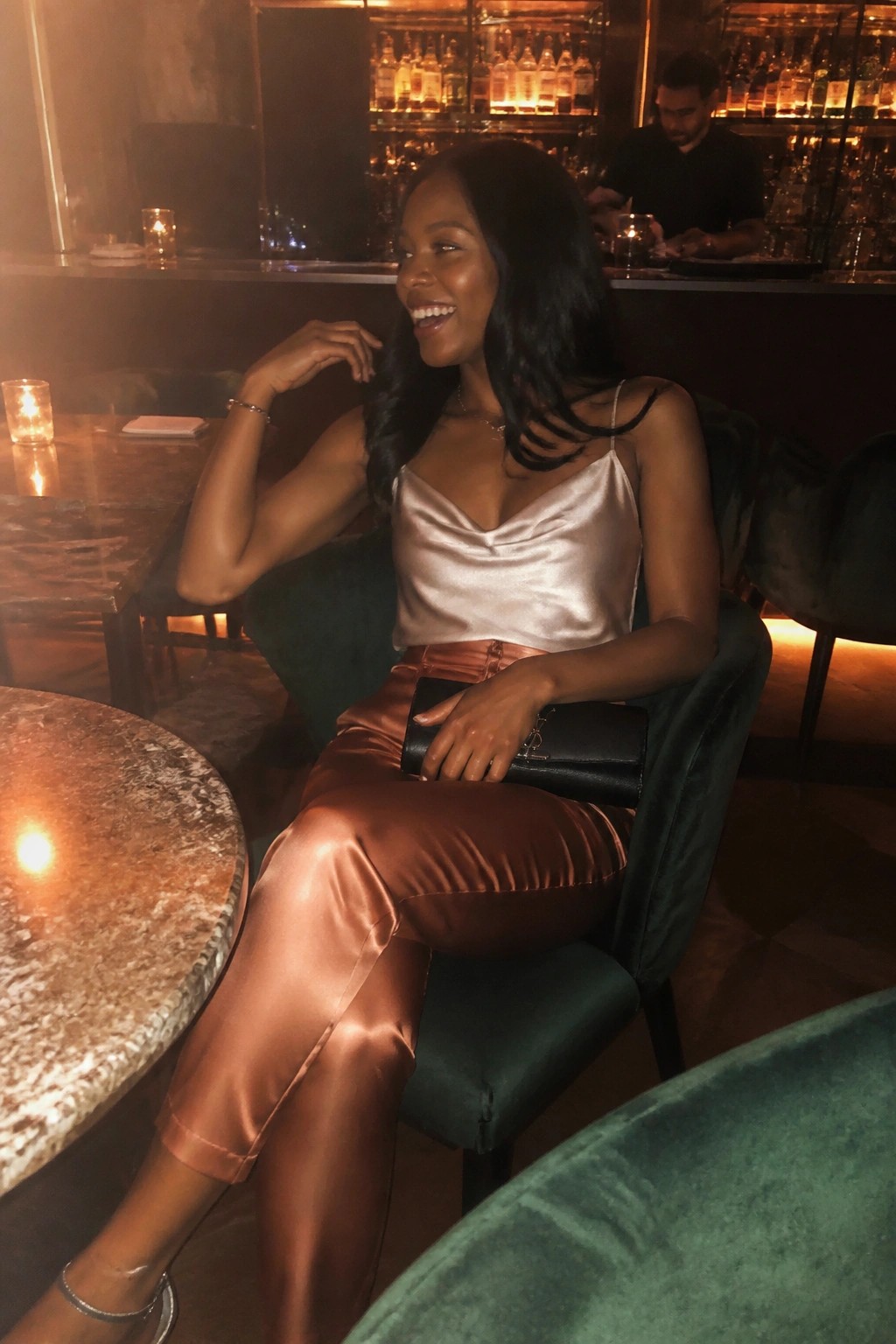 Woman in beige satin top and copper pants seated in green velvet chair at bar