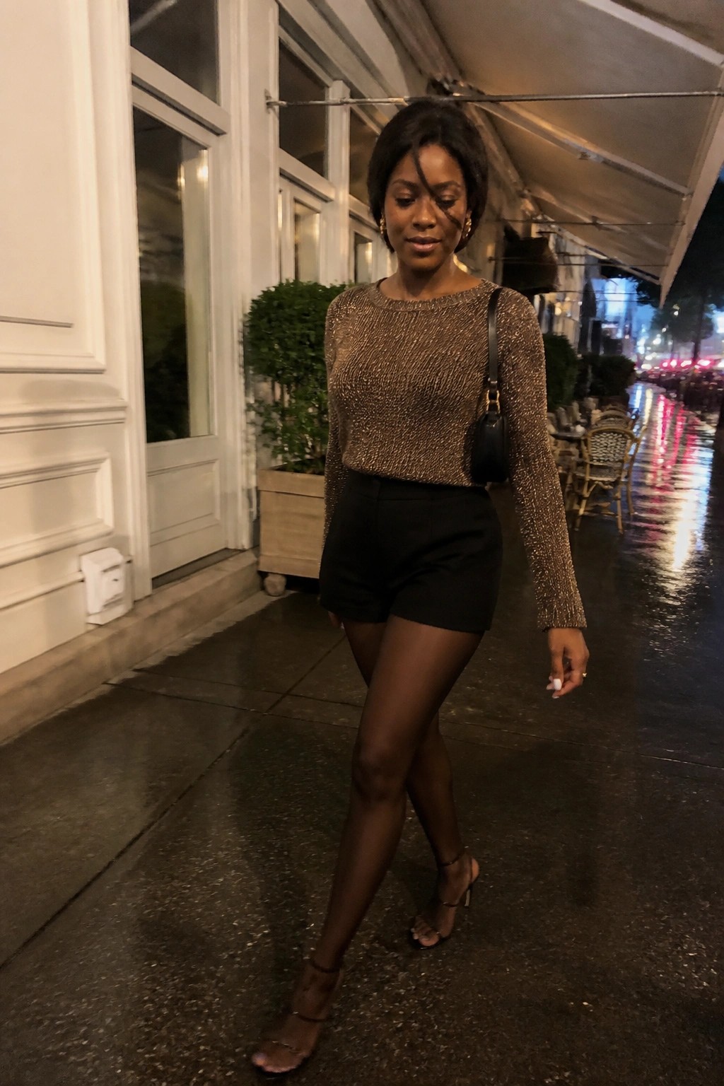 Woman in sparkly gold top and black shorts walks on wet nighttime sidewalk.