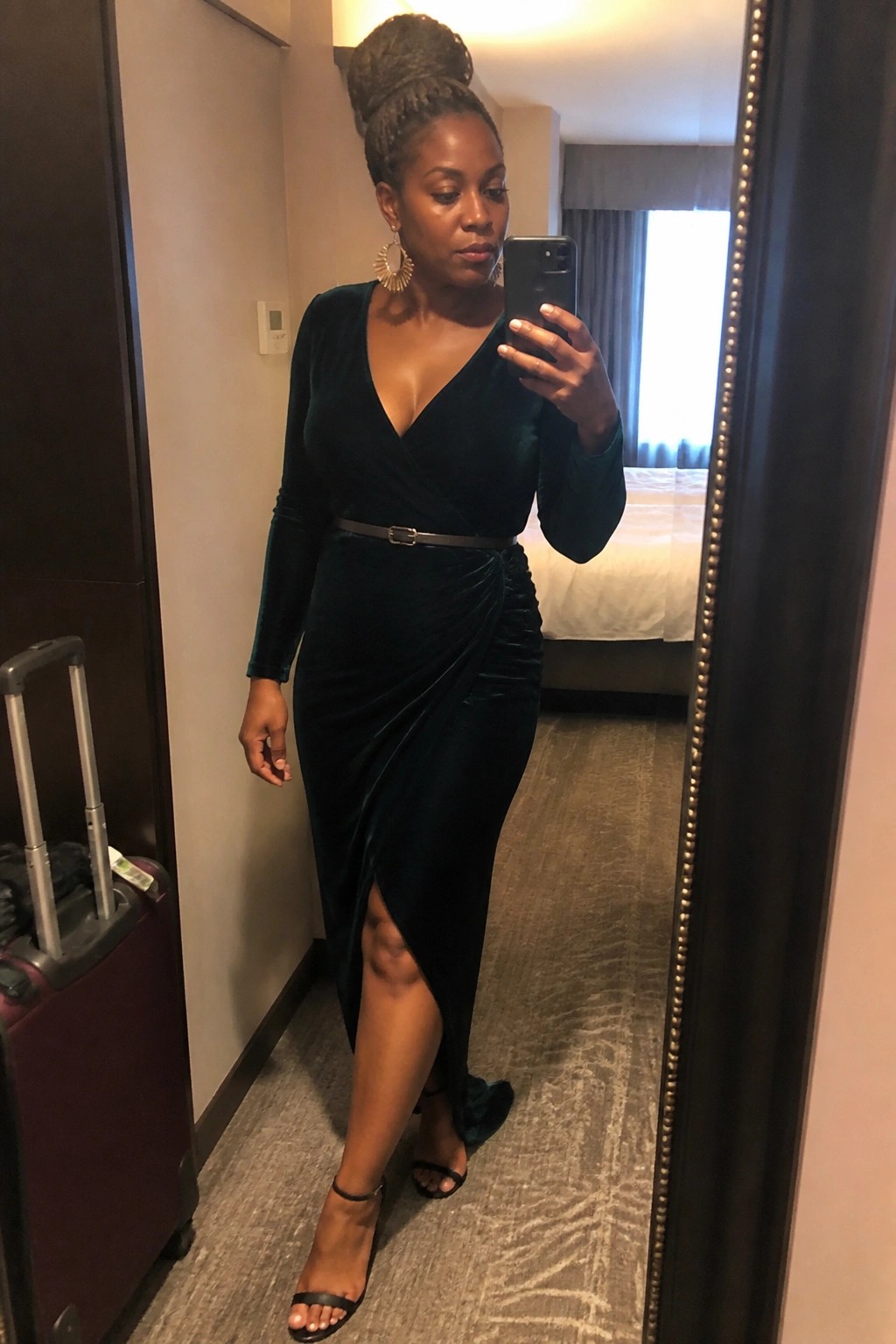 Woman in dark green velvet dress taking mirror selfie in hotel room with suitcase nearby