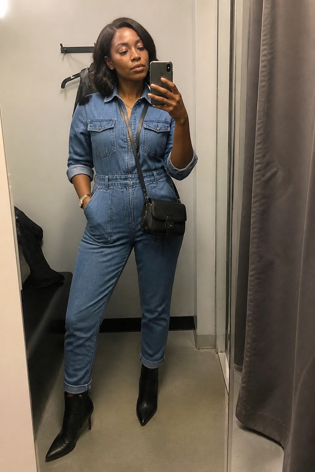 Woman in blue denim jumpsuit and black boots taking mirror selfie in fitting room