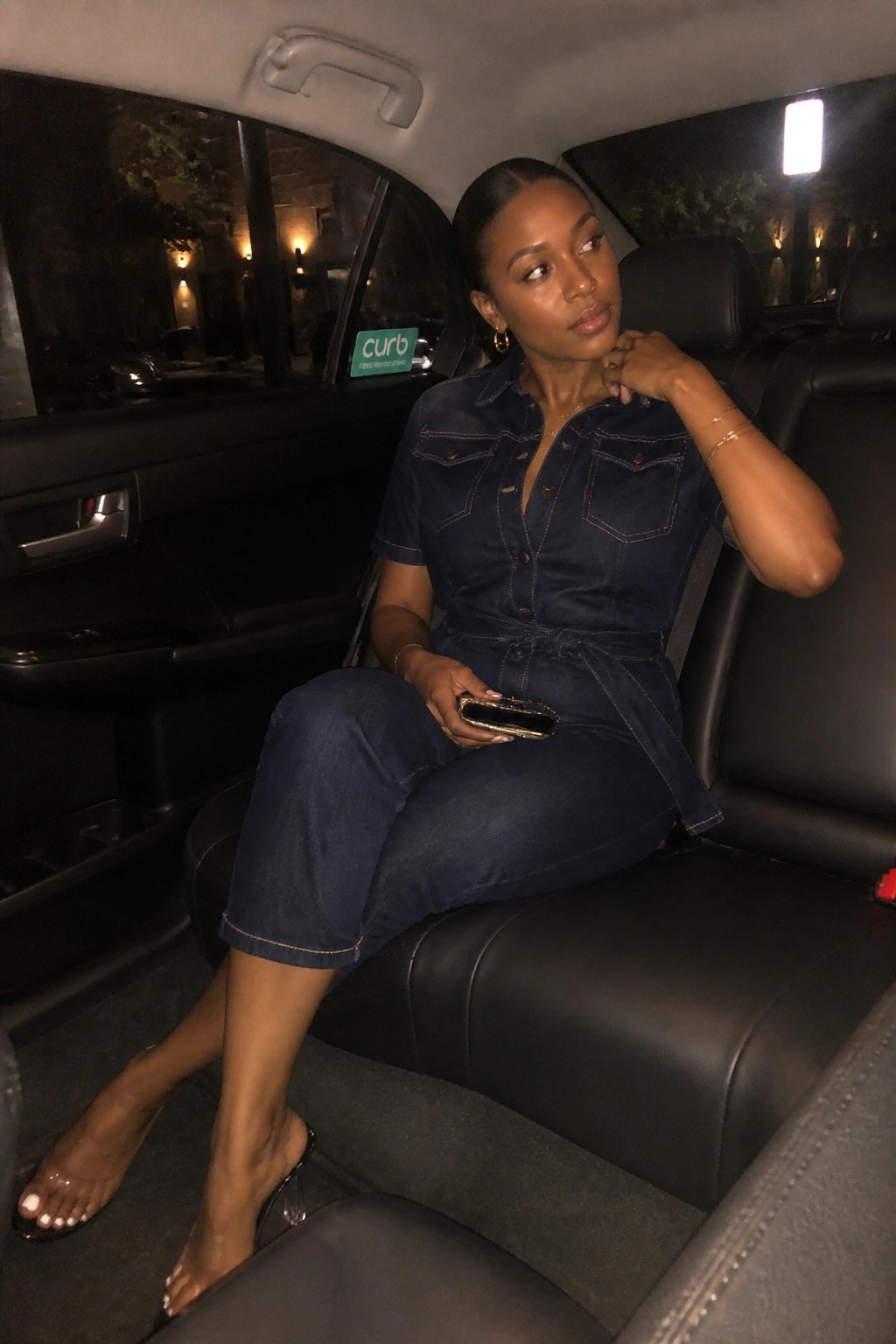 Woman in blue denim jumpsuit seated in car backseat at night holding phone