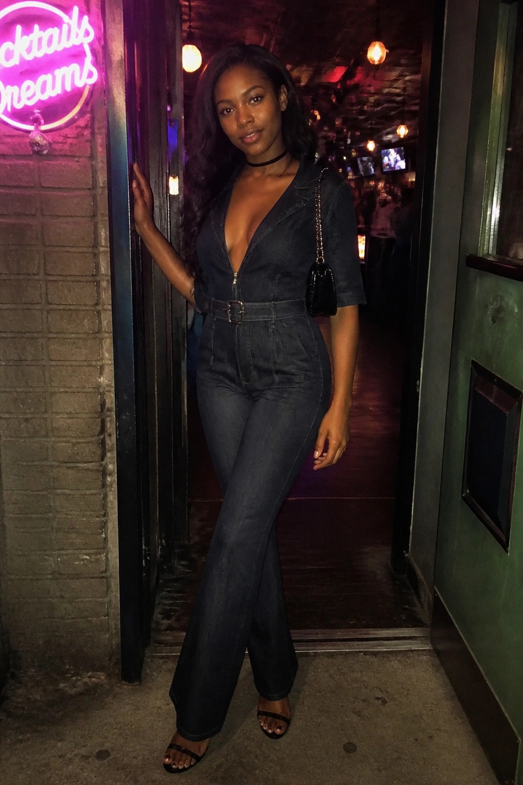 Woman in dark denim jumpsuit with black purse poses at neon bar doorway