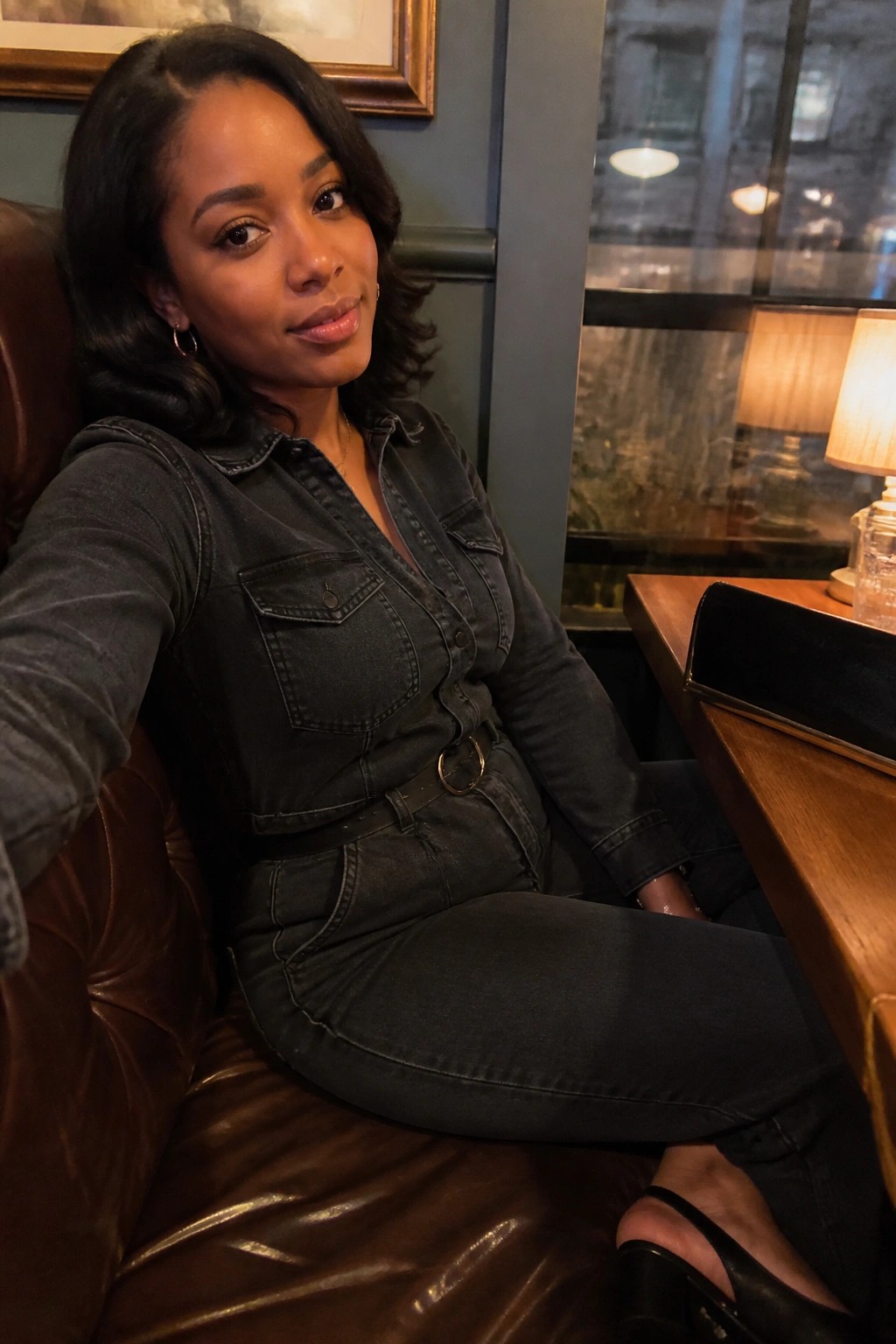 Woman in dark denim jumpsuit seated on brown leather couch in restaurant booth