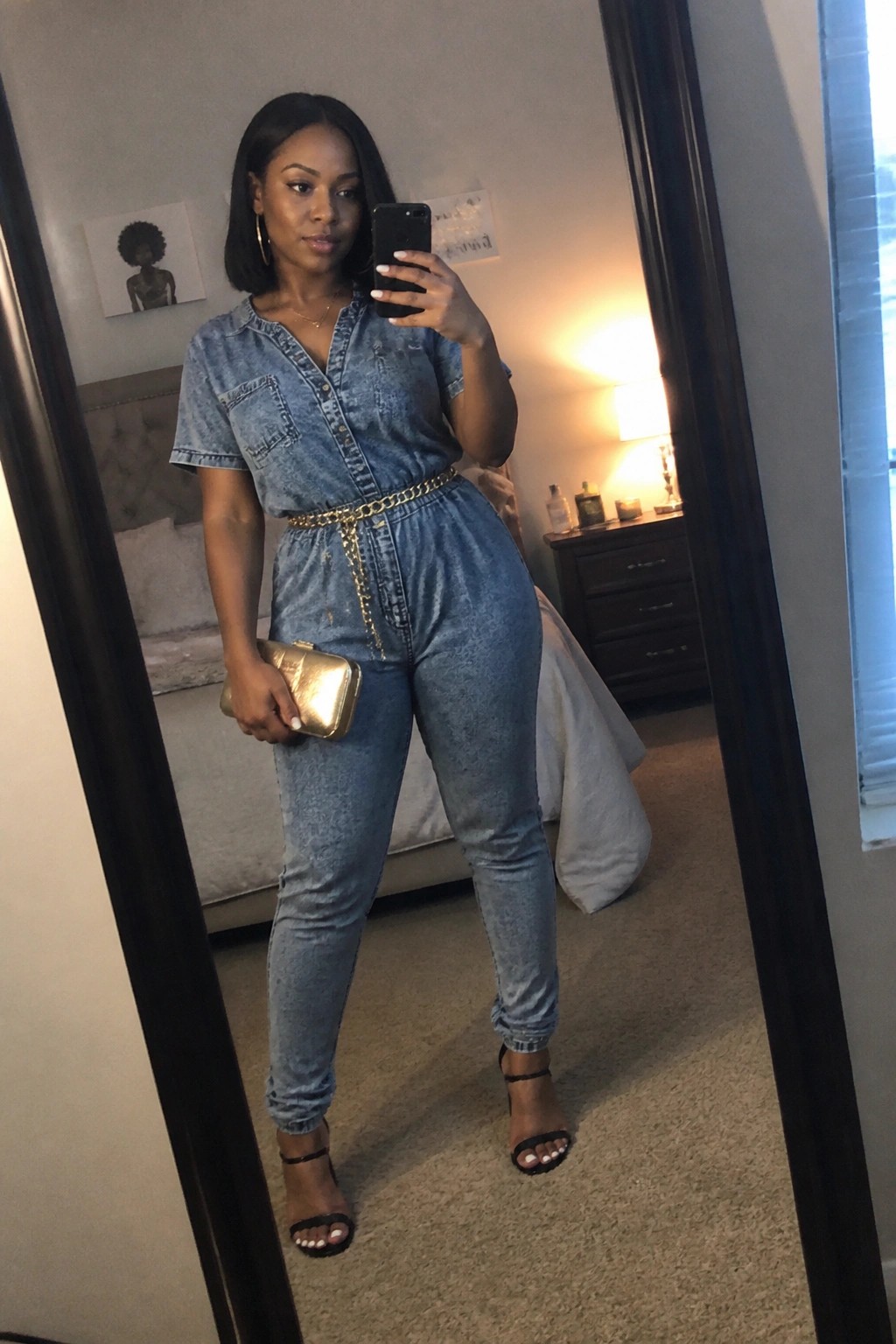 Woman in blue denim jumpsuit with gold chain belt taking bedroom mirror selfie