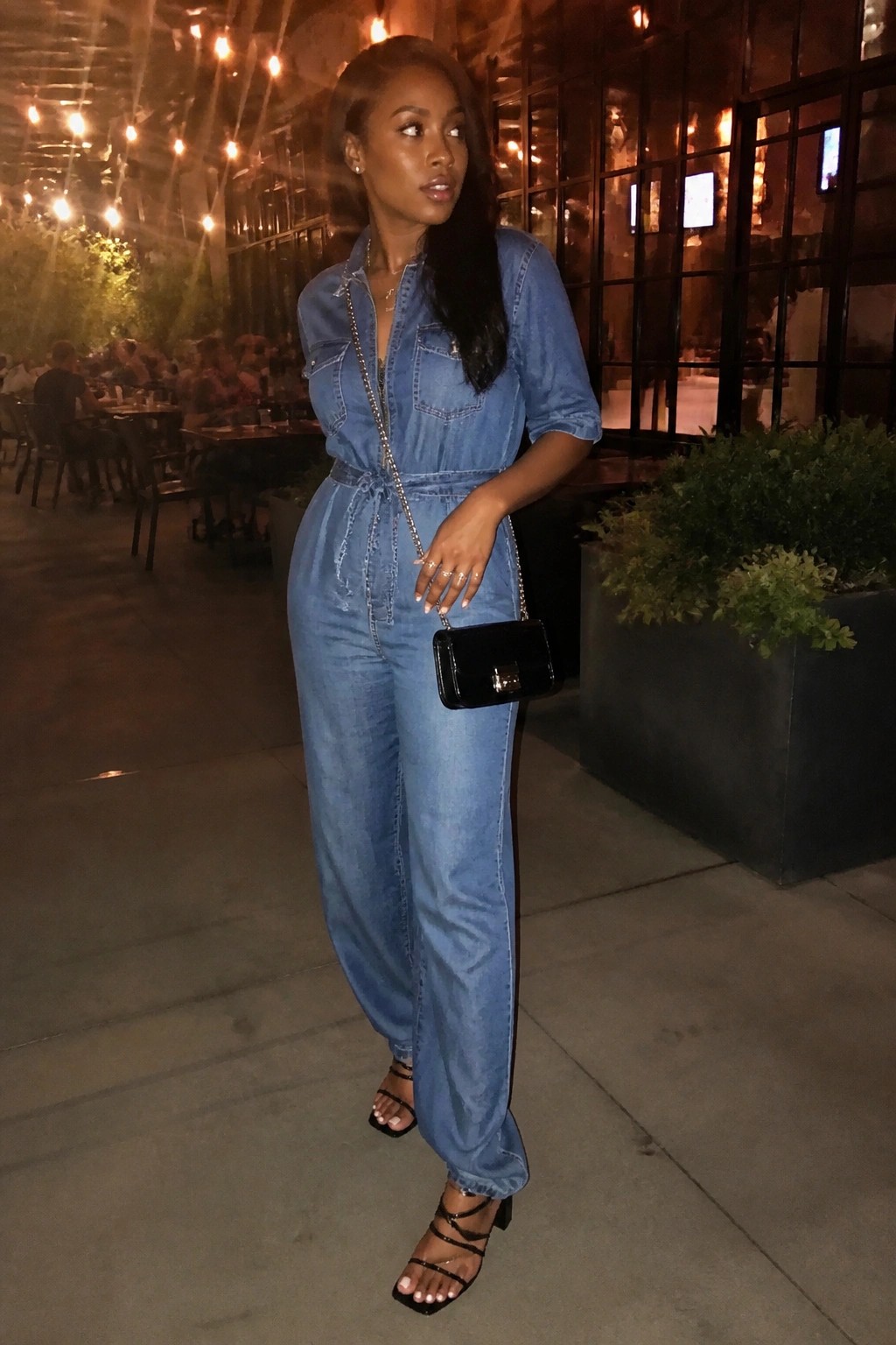 Woman in blue denim jumpsuit with black handbag at night outdoor restaurant