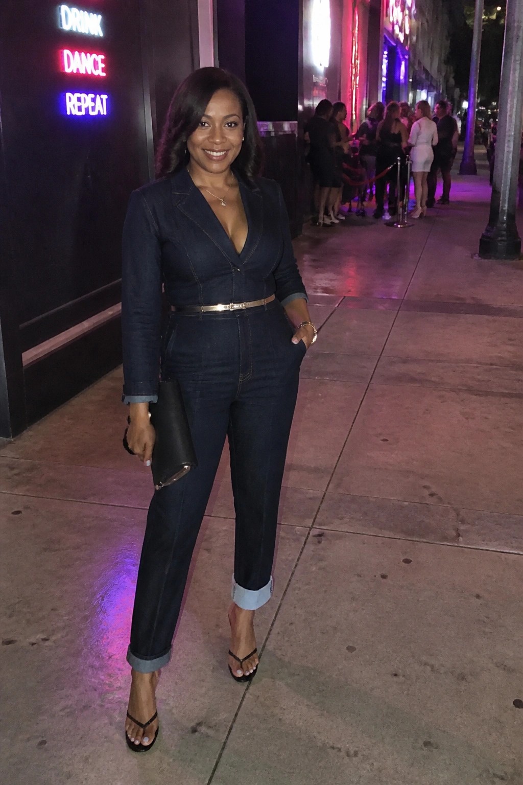 Woman in dark denim jumpsuit with gold belt holds black clutch on neon-lit sidewalk.