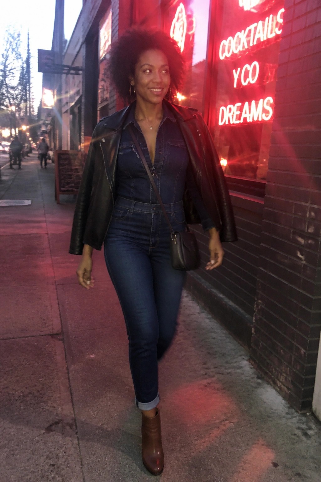Woman in denim jumpsuit and leather jacket walks past glowing red neon sign