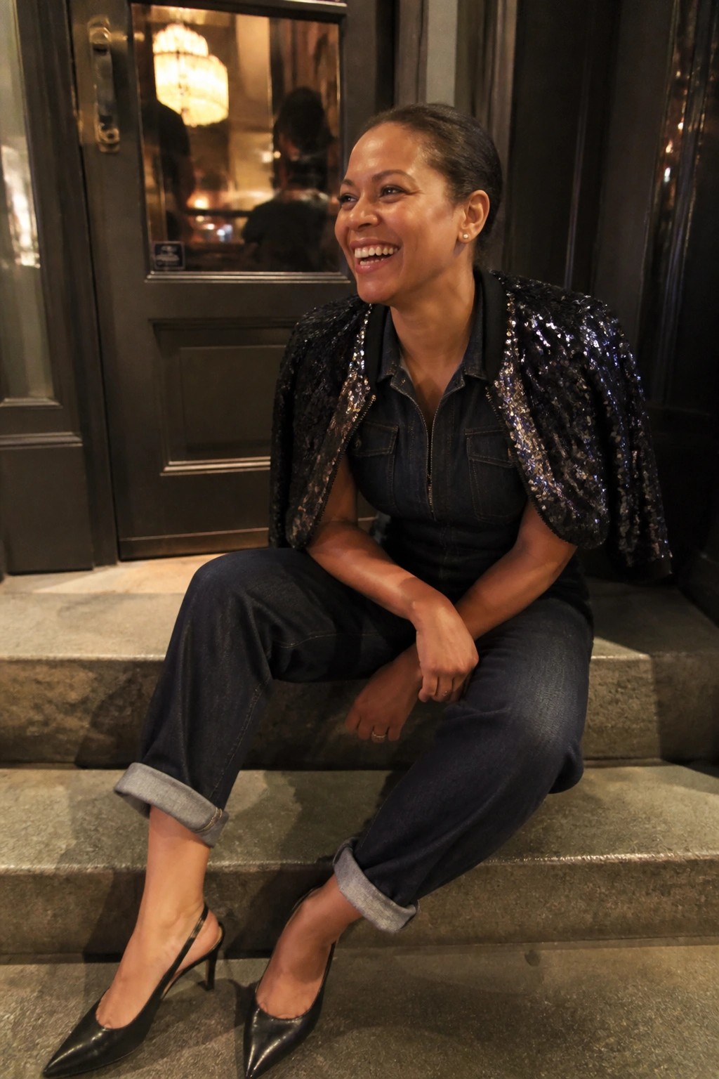 Woman in denim jumpsuit and sequined cape smiling on stone steps outside door