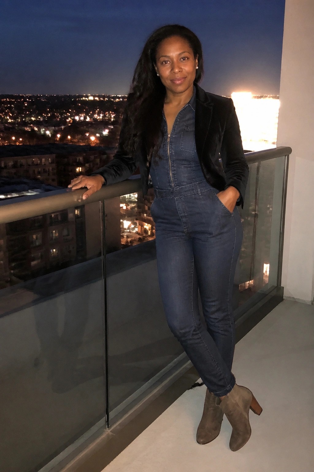 Woman in blue denim jumpsuit and black jacket poses on nighttime city balcony railing