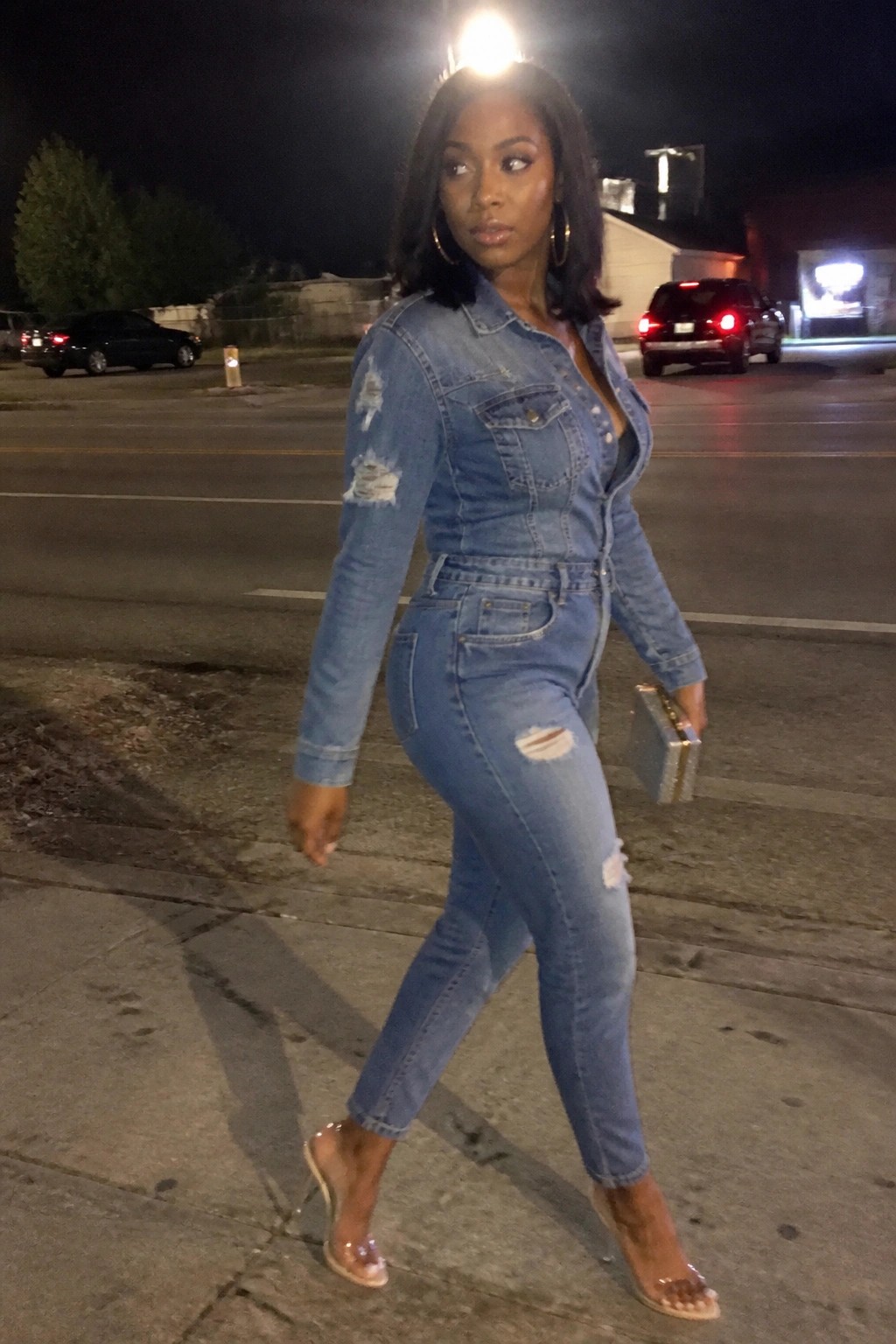 Confident woman in fitted ripped denim jumpsuit and clear heels on city street at night.