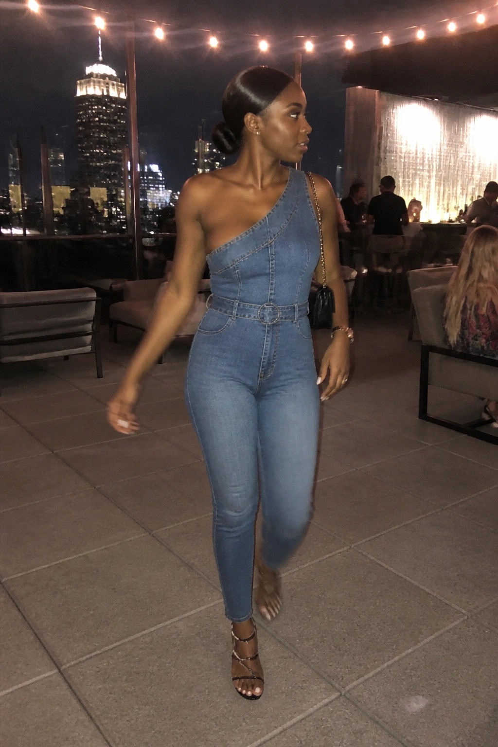 Woman in blue one-shoulder denim jumpsuit on rooftop overlooking city skyline at night.