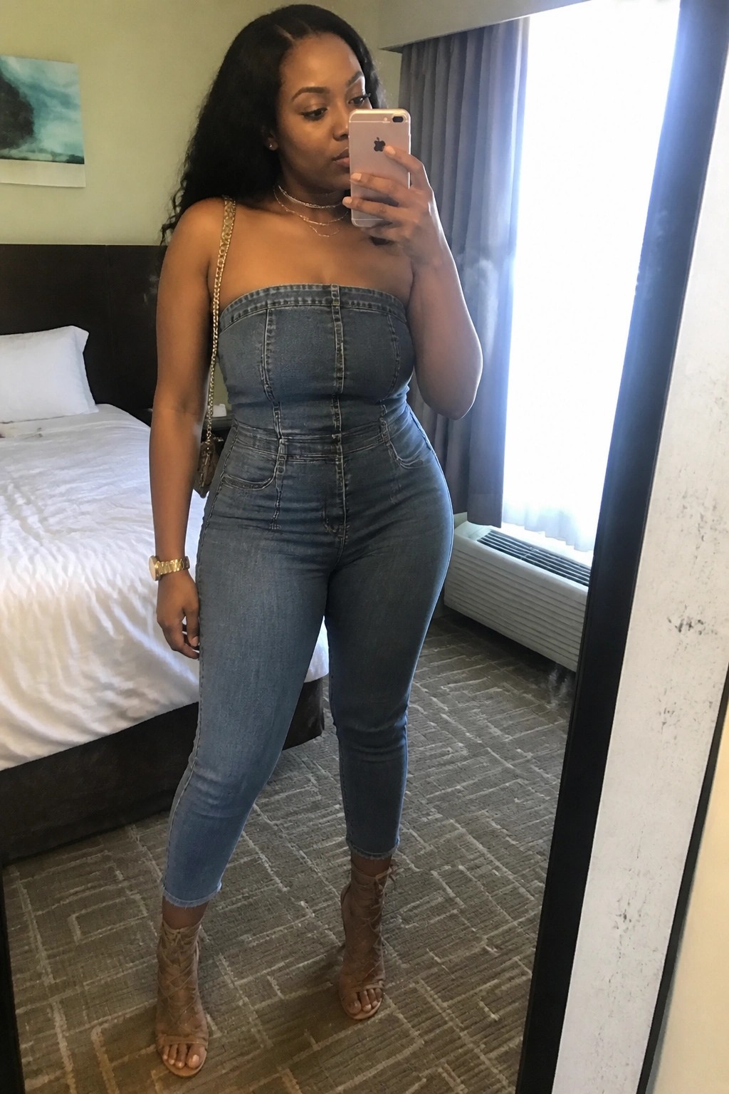 Woman wearing a strapless light blue denim jumpsuit with a gold chain purse strap, layered necklaces, gold watch, and lace up tan heeled booties in a hotel room mirror selfie.