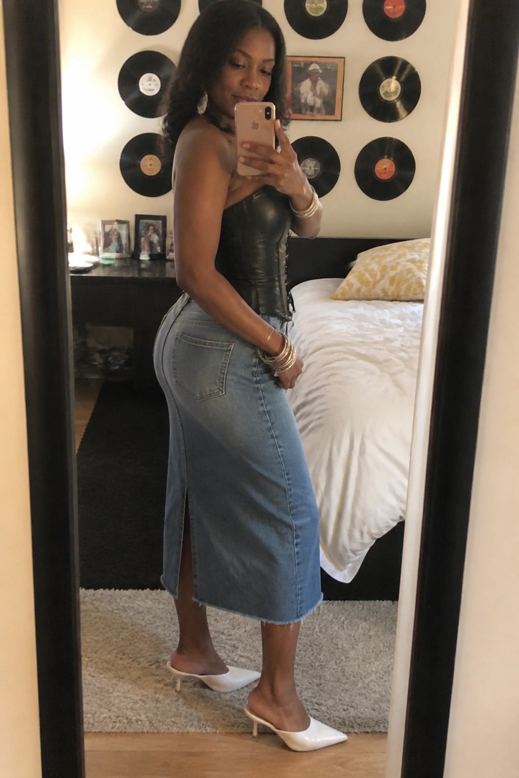 Woman in black leather corset and long denim skirt taking bedroom mirror selfie.