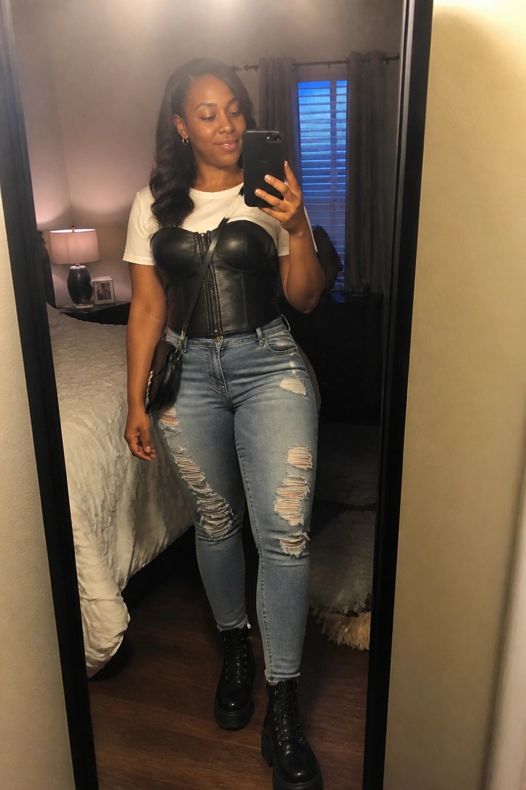 Woman in white short sleeve tee under black leather corset top, light blue ripped skinny jeans, black combat boots, and small black crossbody bag taking mirror selfie in bedroom.