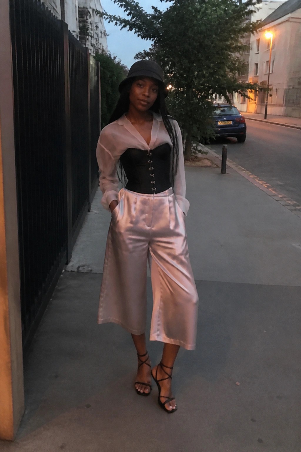 Black woman in black corset over sheer white shirt paired with blush satin wide leg pants black bucket hat and strappy heels standing on sidewalk