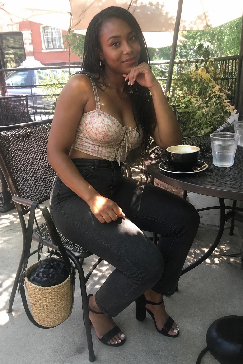 Woman in floral corset crop top black jeans and black block heel sandals with woven bag at outdoor table.