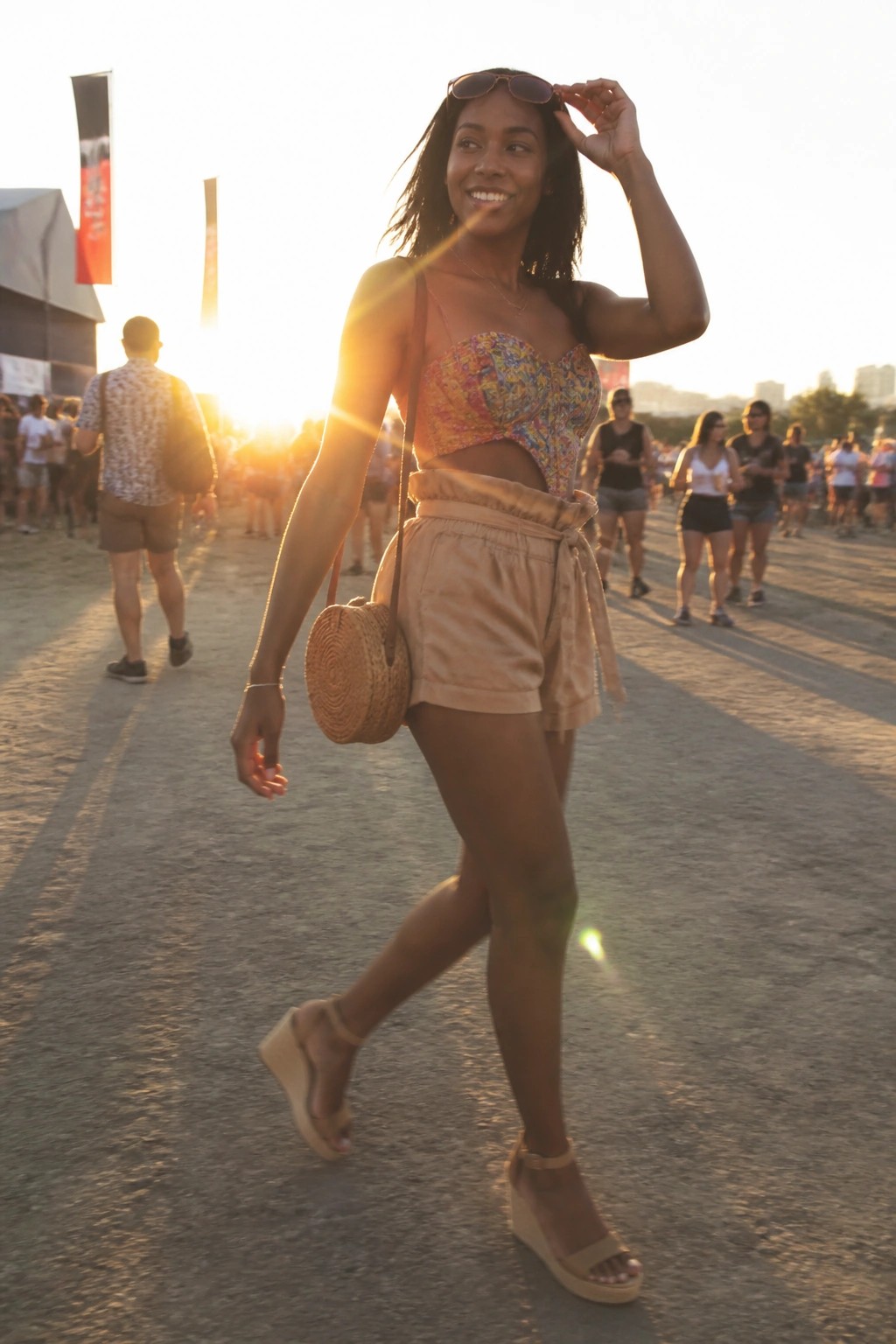 Woman in a colorful floral corset top with cutout waist paired with high-waisted beige shorts tied at the front, round woven crossbody bag, platform wedge sandals, and sunglasses resting on her head.