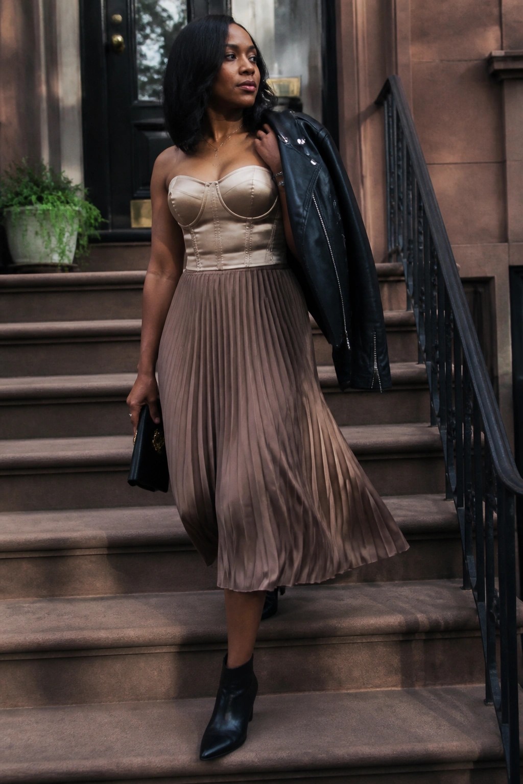 Woman in beige corset top and taupe pleated midi skirt with black leather jacket draped over shoulder, paired with black ankle boots and clutch on stone steps.